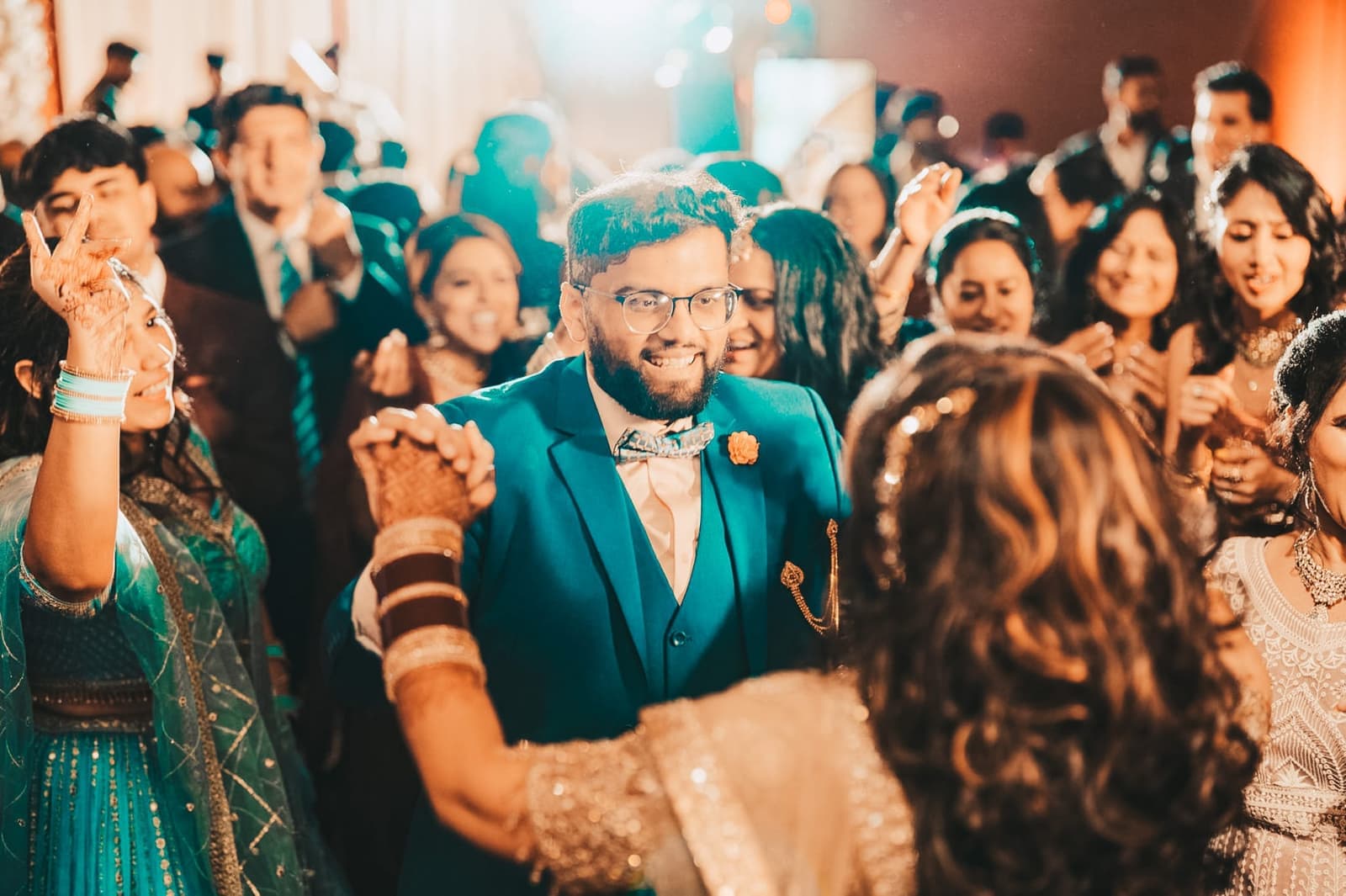 Groom in a teal suit dancing in a warmly lit reception crowd