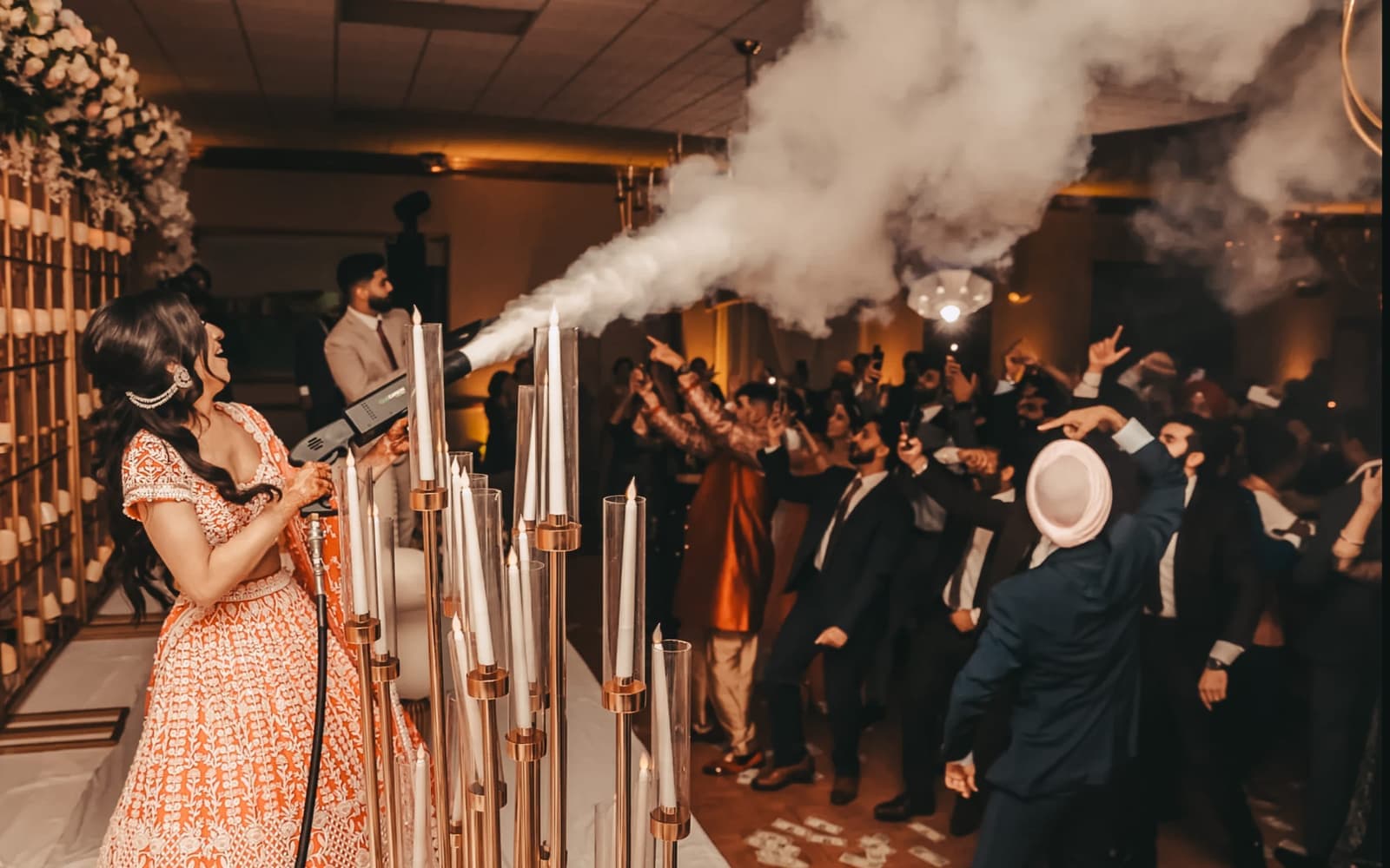 Bride in orange lehenga triggering a cold spark effect over candlesticks as guests cheer