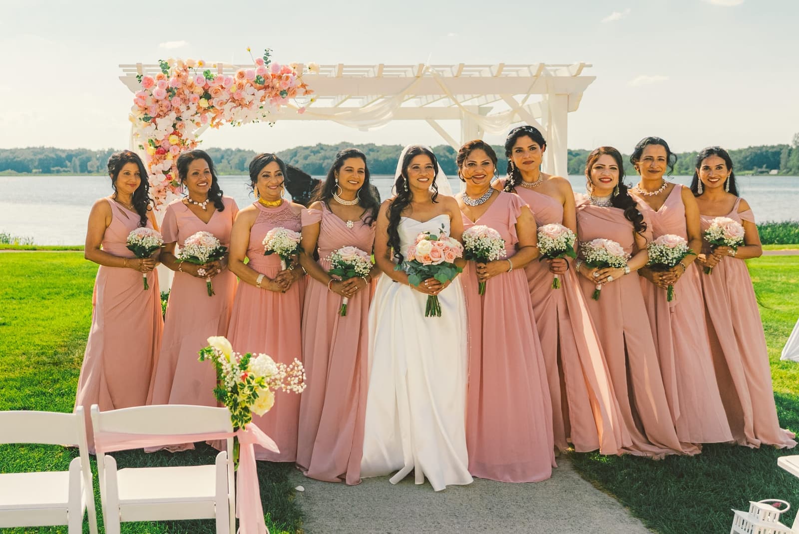 Bride with her bridesmaids in blush gowns beneath a floral arch by a lake
